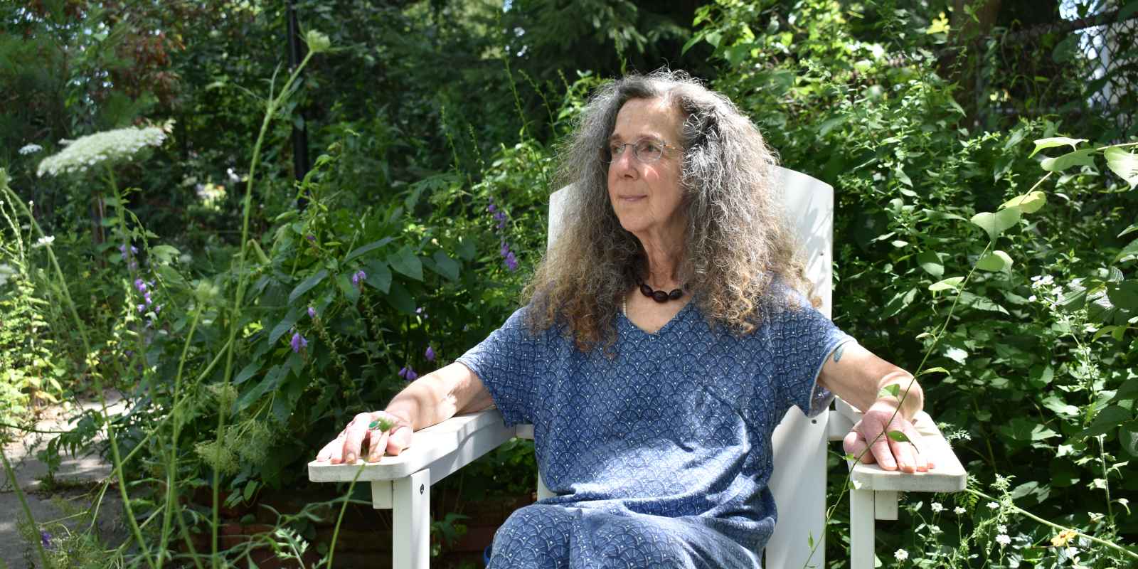 A white woman with curly, graying hair sits in her garden, gazing away from the camera