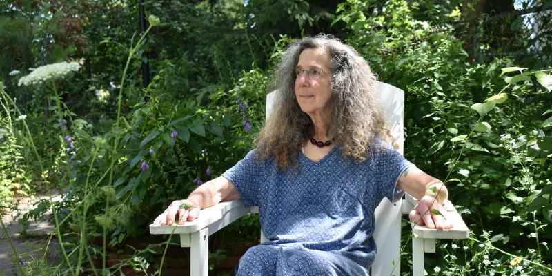 A white woman with curly, graying hair sits in her garden, gazing away from the camera
